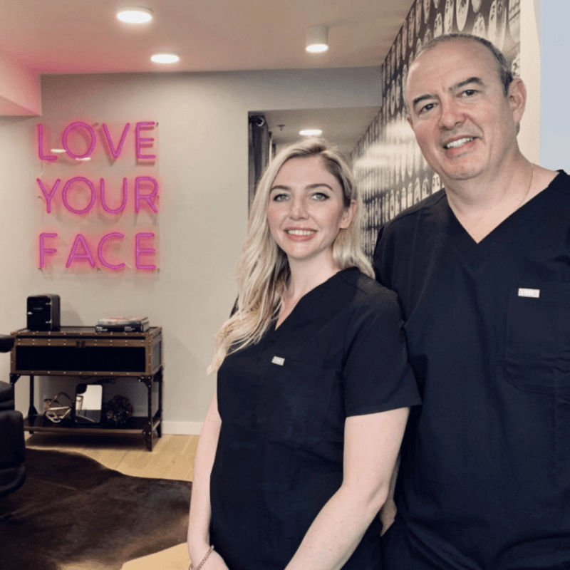 Dr. Oz Fernandez and NP Austin Marie Jacobus standing in black scrubs in front of a sign that reads "love your face"