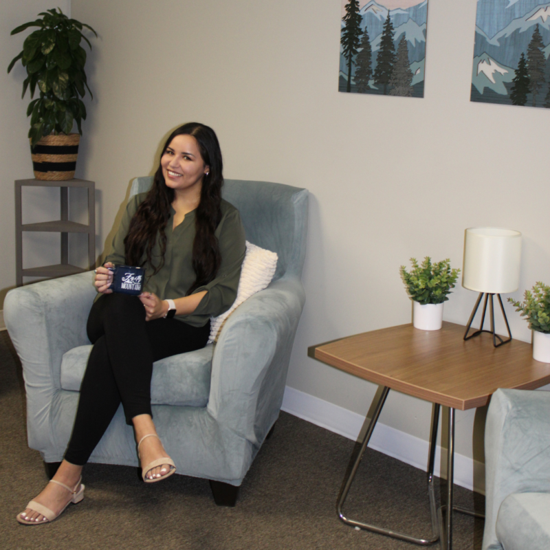 Mountains Therapy Founder Cristina Andino, LCSW, MSW sitting in a blue chair next to a coffee table holding a mug