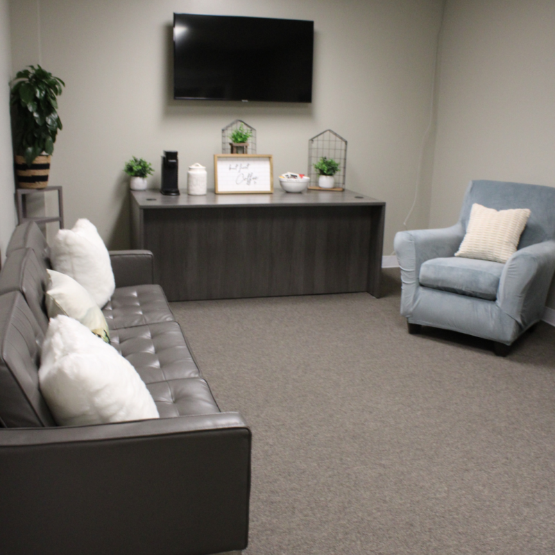 Mountains Therapy treatment room with black couch and white pillows, a blue chair, a table against the wall, and a tv on the wall
