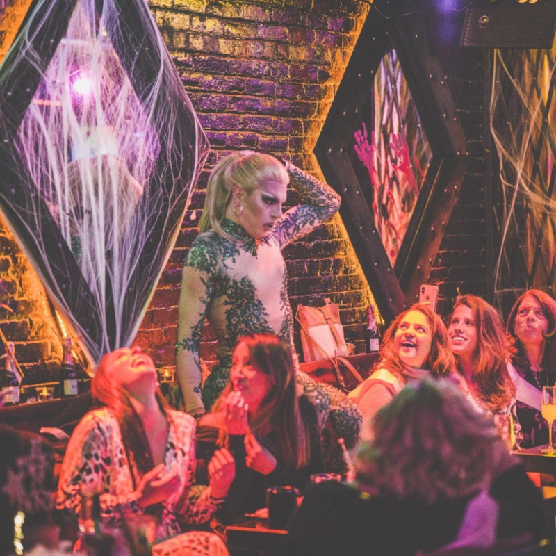 Drag Performer with blonde hair in ponytail standing behind a group of women sitting at a table