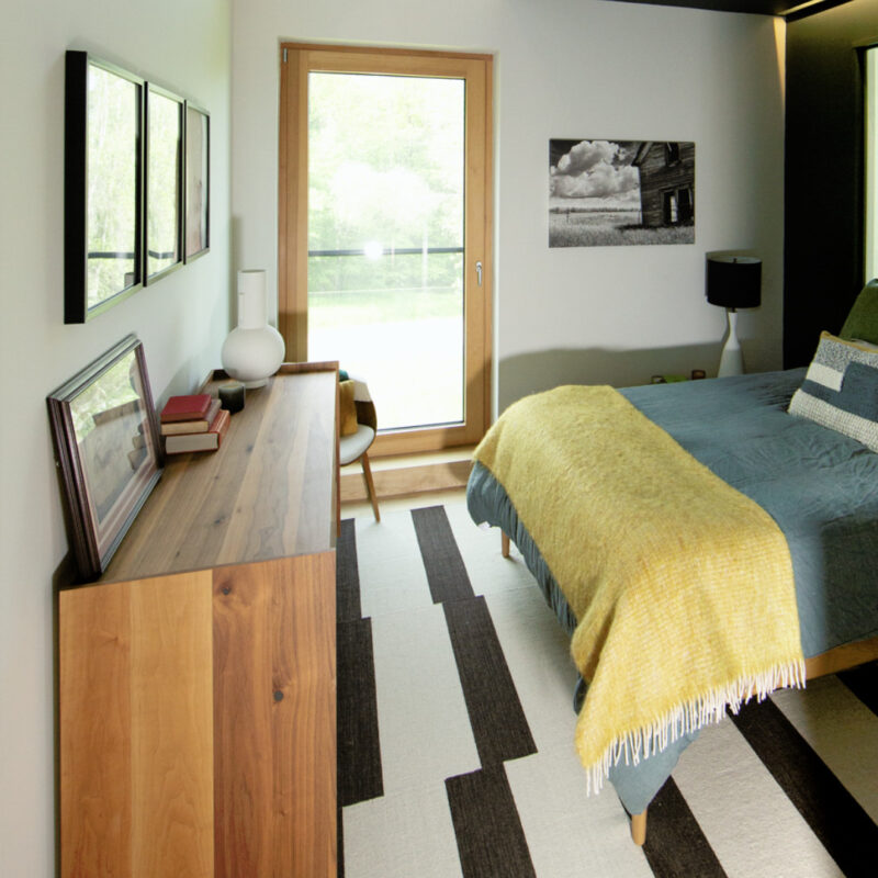 Bedroom in The Catskill Project home with black and white rug, wooden dresser, bed, and large window