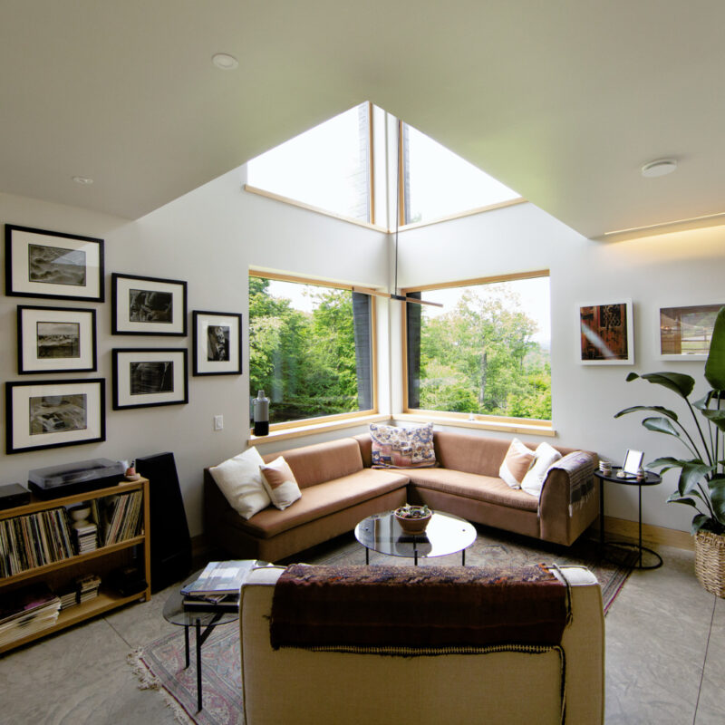 Cozy living room in The Catskill Project home with large windows letting in natural light and views of trees, a corner couch. yellow chair, and framed pictures on the wall