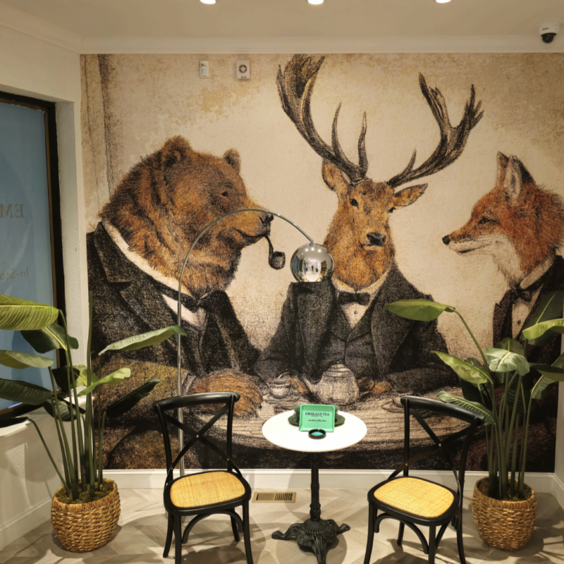 Wall painting featuring a bear, deer, and fox wearing suits on the wall behind a small table with two chairs and two plants 