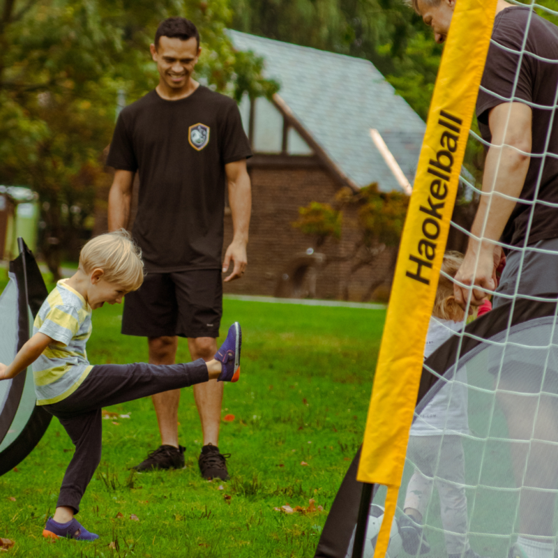 A kid kicking a soccer ball into a goal with two coaches nearby