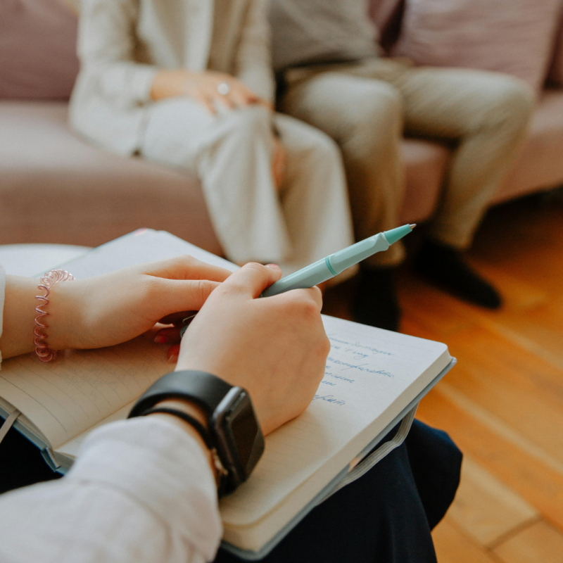 Hands holding a pen resting on a notebook and two people sitting on a couch